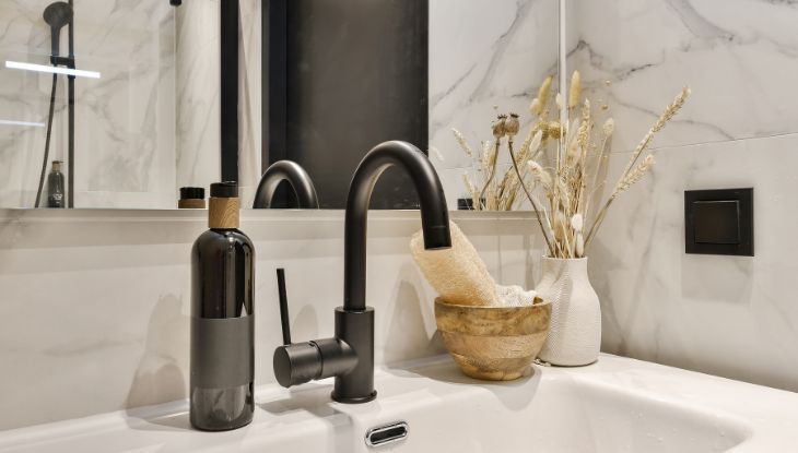 Moderno kupatilo sa mat crnom slavinom, dekorativnim bocama i suvim aranžmanom na beloj mermernoj pozadini.